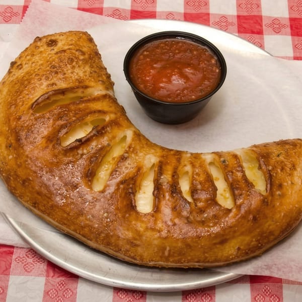 A golden-brown calzone with visible cheese filling, served with a side of marinara sauce on a checkered tablecloth.