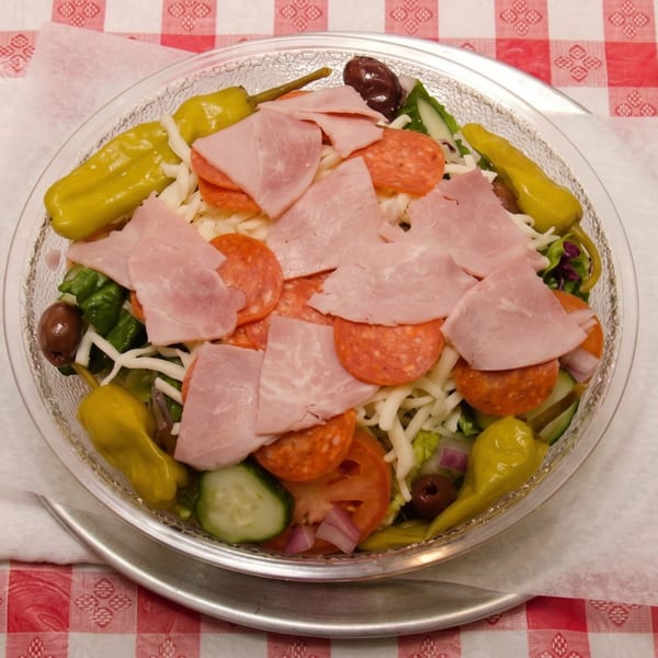 A large antipasto salad with ham, pepperoni, cheese, and vegetables in a clear bowl on a red checkered tablecloth.
