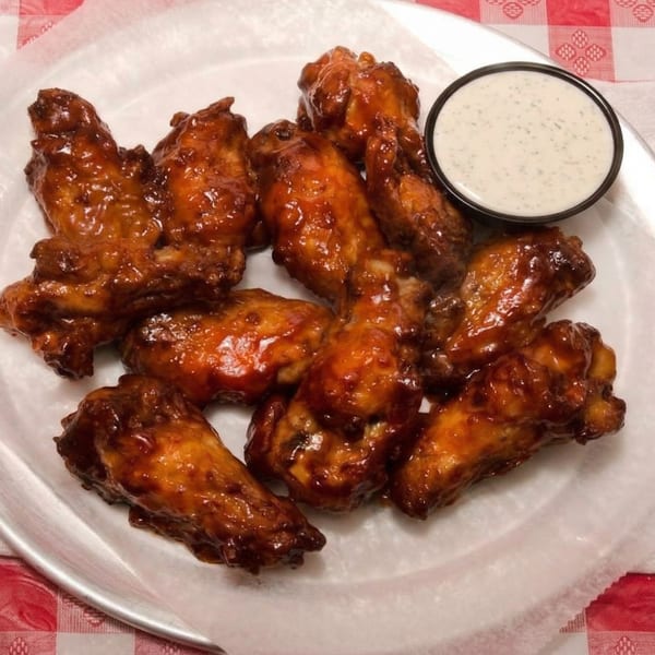 A plate of glistening, saucy BBQ chicken wings with a side of ranch dressing on a red and white checkered tablecloth.